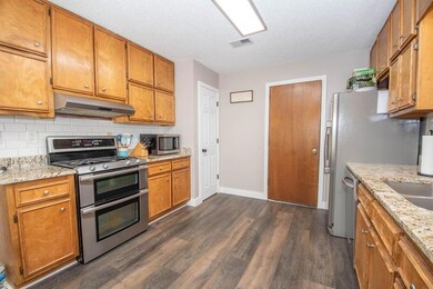 Kitchen with appliances with stainless steel finishes, under cabinet range hood, dark wood-style flooring, decorative backsplash, and brown cabinetry