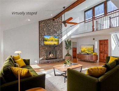 Living area featuring high vaulted ceiling, wood finished floors, a ceiling fan, a stone fireplace, and beam ceiling