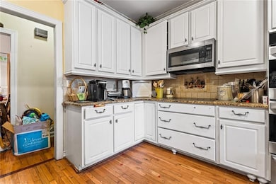 Kitchen featuring stainless steel microwave, white cabinetry, tasteful backsplash, light wood finished floors, and stone countertops