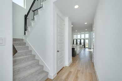 Bright and modern entryway with light wood-like ceramic flooring and white walls. It features a carpeted staircase with a black railing leading upstairs. The hallway opens into a spacious living area with large windows, offering plenty of natural light.