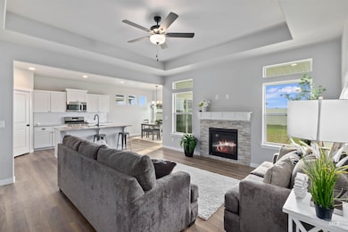 Living area featuring a raised ceiling, dark wood-style flooring, ceiling fan, and a stone fireplace