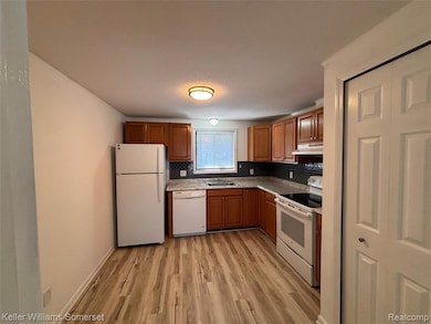 Kitchen featuring white appliances, brown cabinetry, backsplash, light wood-style flooring, and under cabinet range hood