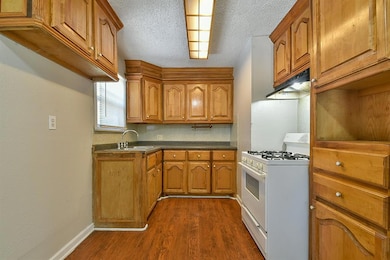 Kitchen featuring white range with gas cooktop, brown cabinetry, dark wood-style flooring, under cabinet range hood, and dark countertops