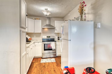 Kitchen featuring white appliances, wall chimney exhaust hood, light wood-type flooring, white cabinetry, and a textured ceiling