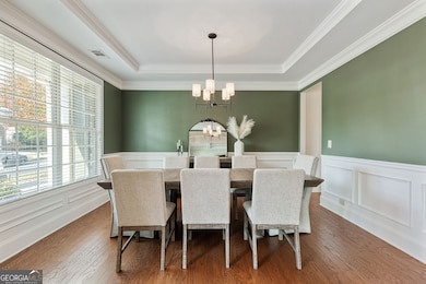 This elegant dining room features soft sage-green walls with crisp white wainscoting, rich hardwood floors, and a stylish chandelier that adds a touch of sophistication. Large windows fill the space with natural light, creating a warm and inviting se