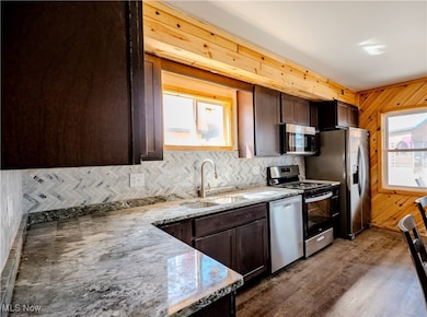 Kitchen with dark brown cabinets, wooden walls, appliances with stainless steel finishes, decorative backsplash, and light stone counters