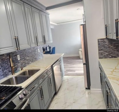 Kitchen featuring backsplash, gray cabinetry, light marble finish floors, light stone countertops, and appliances with stainless steel finishes