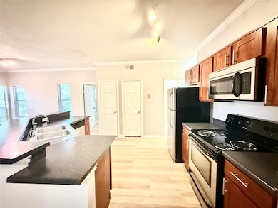 Kitchen with appliances with stainless steel finishes, light wood finished floors, dark countertops, crown molding, and an island with sink