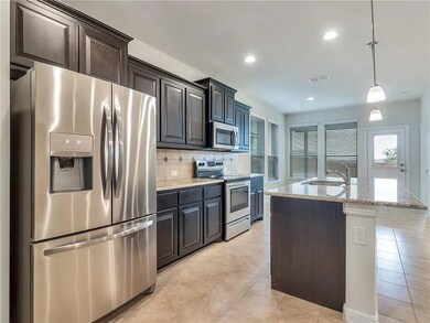 Kitchen with stainless steel appliances, backsplash, light stone countertops, hanging light fixtures, and a kitchen island with sink