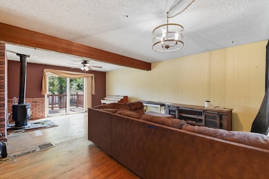 Living room featuring a wood stove, wood finished floors, a textured ceiling, beam ceiling, and a ceiling fan