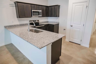 Kitchen featuring dark brown cabinets, light stone countertops, stainless steel appliances, a peninsula, and concrete flooring