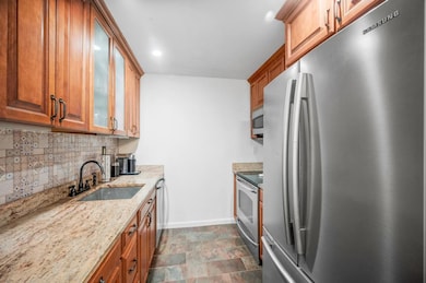 Kitchen with appliances with stainless steel finishes, tasteful backsplash, light stone counters, brown cabinetry, and glass insert cabinets