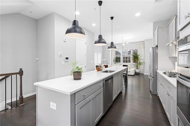 Kitchen featuring appliances with stainless steel finishes, gray cabinetry, an island with sink, dark wood-type flooring, and light countertops