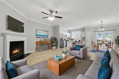 Living room with crown molding, wood finished floors, a warm lit fireplace, a chandelier, and a ceiling fan