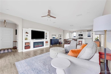 Living area with a ceiling fan, light wood-style flooring, arched walkways, and a glass covered fireplace