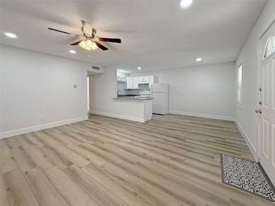 Bright and open living space with modern laminate flooring, recessed lighting, and a ceiling fan. The kitchen features white cabinetry, granite countertops, and essential appliances. The room is spacious and inviting, ideal for both relaxing and entertaining.
