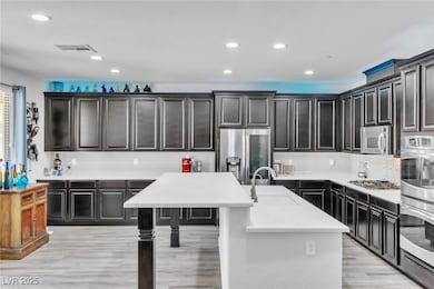 Kitchen featuring recessed lighting, appliances with stainless steel finishes, an island with sink, dark cabinets, and light wood-style flooring
