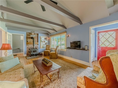 Living area with a wood stove, beamed ceiling, wood finished floors, and ceiling fan