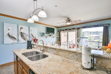 This cozy living space features an open kitchen with wooden cabinets and a breakfast bar with stools. The room has a ceiling fan, tiled floors, and light blue walls. A small dining area with a round table and two chairs sits beneath wall-mounted art and a TV. Perfect for relaxed, comfortable living.