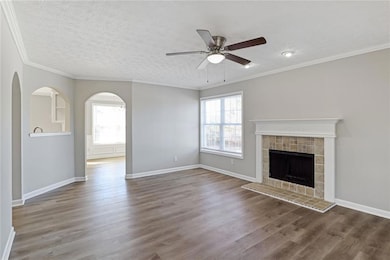 Unfurnished living room with a ceiling fan, wood finished floors, a tiled fireplace, crown molding, and arched walkways