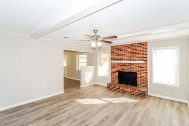 Unfurnished living room featuring wood finished floors, beamed ceiling, a brick fireplace, ceiling fan, and crown molding