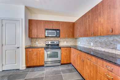 Kitchen featuring backsplash, dark stone counters, stainless steel appliances, brown cabinets, and dark tile patterned flooring