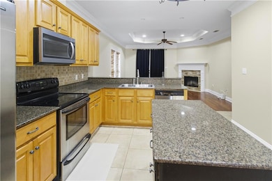 Kitchen featuring appliances with stainless steel finishes, ornamental molding, a tray ceiling, a stone fireplace, and dark stone counters