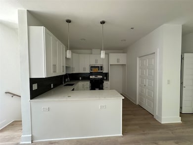 This photo showcases a modern kitchen with white cabinetry, a central island with a marble countertop, and pendant lighting. The space features a sleek black backsplash, stainless steel appliances, and wood flooring, offering a clean and contemporary look.