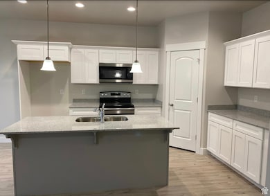 Kitchen with white cabinetry, light stone countertops, stainless steel appliances, a center island with sink, and decorative light fixtures