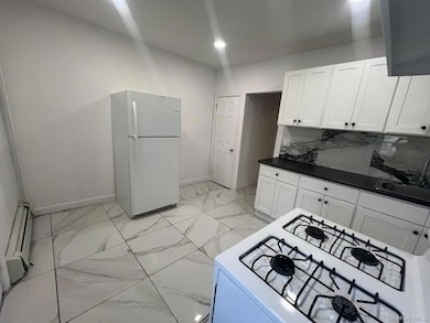 Kitchen featuring dark countertops, white appliances, light marble finish flooring, baseboard heating, and white cabinets