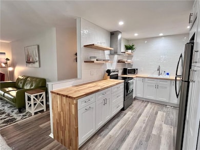 Kitchen with butcher block counters, open shelves, stainless steel appliances, wall chimney exhaust hood, and tasteful backsplash