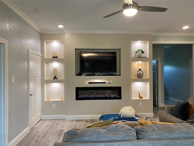 Living area with built in shelves, crown molding, light wood-style floors, a glass covered fireplace, and a ceiling fan