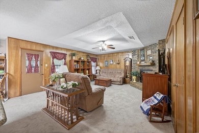 Carpeted living area with wooden walls, ceiling fan, a textured ceiling, a raised ceiling, and a brick fireplace