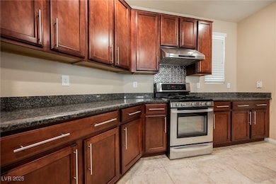 Kitchen featuring stainless steel range with gas cooktop, under cabinet range hood, dark stone countertops, backsplash, and light tile patterned floors