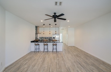 Modern, open concept living space featuring a sleek kitchen with a breakfast bar and three pendant lights. The room has vinyl-style flooring and a ceiling fan, creating a bright, airy atmosphere.