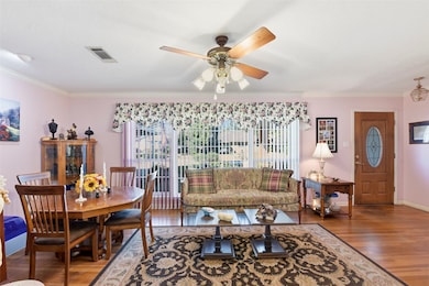 This inviting living and dining area features hardwood floors, a ceiling fan, and large windows with floral valances offering plenty of natural light. The space includes a cozy sofa, a classic dining set, and decorative accents, creating a warm, welcoming atmosphere.