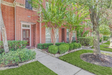 A private pathway framed by lush, professionally maintained hedges and stone-edged flowerbeds leads to the stately front door. Mature crepe myrtles and manicured greenery line the sidewalk, offering shade, privacy, and beautiful curb appeal that complements the home’s striking red brick facade in this peaceful, tree-lined section of Rice Military.
