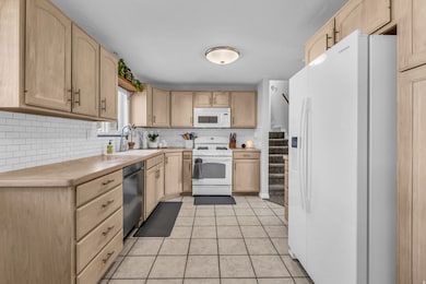 Kitchen with white appliances, light brown cabinetry, backsplash, light tile patterned flooring, and light countertops