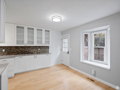 Kitchen with white cabinets, tasteful backsplash, glass insert cabinets, light wood finished floors, and light stone counters