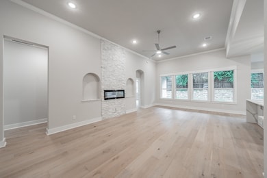 Unfurnished living room featuring ornamental molding, light wood-type flooring, a stone fireplace, ceiling fan, and arched walkways