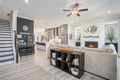 Living room featuring stairs, a ceiling fan, a glass covered fireplace, light wood-style floors, and ornamental molding
