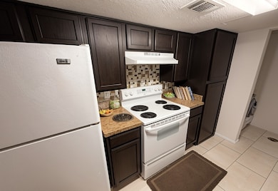 Kitchen featuring white appliances, dark brown cabinetry, backsplash, light tile patterned floors, and under cabinet range hood