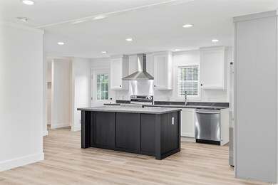 Kitchen with wall chimney range hood, appliances with stainless steel finishes, a kitchen island, white cabinets, and light wood-type flooring