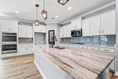 Kitchen with ornamental molding, appliances with stainless steel finishes, hanging light fixtures, light stone counters, and a large center island with sink.