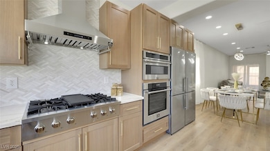 Kitchen featuring open floor plan, light brown cabinets, wall chimney range hood, stainless steel appliances, and light wood finished floors