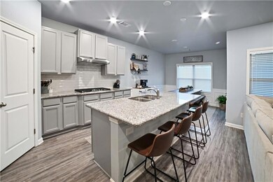 Kitchen featuring light stone counters, gray cabinets, dark wood finished floors, a center island with sink, and tasteful backsplash