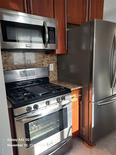 Kitchen with appliances with stainless steel finishes, tasteful backsplash, brown cabinets, light stone counters, and light tile patterned floors