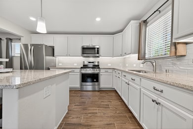 This kitchen features modern stainless-steel appliances, white cabinetry, and granite countertops. The space is well-lit with a large window and recessed lighting, complemented by wood-look tile flooring for a warm, inviting feel.