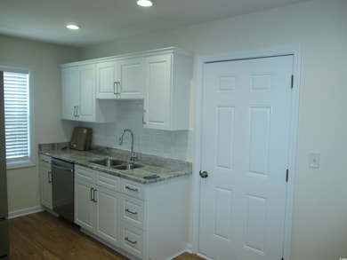 Kitchen featuring white cabinets, decorative backsplash, stainless steel dishwasher, light stone countertops, and dark wood-style floors