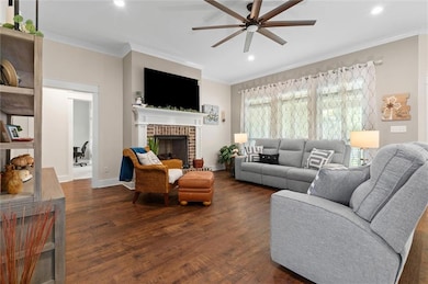 Living area with a ceiling fan, dark wood finished floors, ornamental molding, recessed lighting, and a brick fireplace